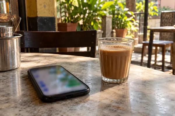 A woman's hands at a cafe table holding a phone, the chat screen a soft blur, a glass of cutting chai beside it. Hands o