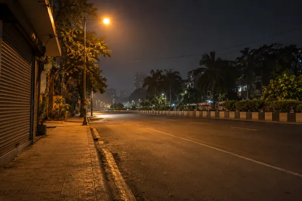 A wide Navi Mumbai road at night from the kerb: a closed shop shutter, one sodium lamp, a parked cab with its rear door